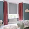 Floor-length pleated mauve curtains flank a lowered grey roller blind, framing and shading a bay window in a teal-walled bedroom with white floors, radiator, bed, and greenery visible outside.