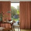 Rust-colored pleated, floor-length curtains, parted at center to reveal sliding glass door; curtains filter warm light. Wooden dining table set for breakfast faces a sunlit backyard garden.