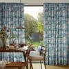 Full-length pleated curtains with blue-green watercolor pattern, parted to frame a sliding door; allowing morning light into a dining nook with wooden table, pendant lamp, breakfast plates, and garden view.