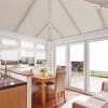 Cream pleated cellular blinds covering triangular conservatory roof panels and full-height windows/doors, drawn closed and diffusing soft daylight into a bright dining-kitchen conservatory with wooden table, chairs and patio view.