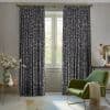 Patterned navy-grey floral drapery panels on a brass rod, drawn partly aside to frame a tall paneled French window, filtering soft daylight in a neutral, furnished sitting room.