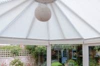 White pleated conservatory blinds, radial and scalloped, cover the roof panels and upper windows, closed and diffusing daylight; bright conservatory interior with a woven spherical pendant and garden view.