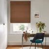 Brown Roman fabric blind, lowered over the window with small folds; filtering soft daylight into a neutral study with a desk, teal chair, vase of flowers and a tree view.