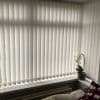 White vertical blinds with subtle leaf pattern, fully closed over an L-shaped bay window, filtering soft daylight; windowsill holds a potted white orchid, radiator below, blanket and cushion nearby.