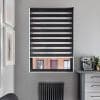 Black zebra roller blind, lowered with alternating opaque and sheer horizontal stripes filtering light; modern grey kitchen corner, radiator below, countertop with espresso machine, framed art and brick-building view.