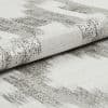 Folded woven fabric with silver metallic horizontal stripes, tightly textured and slightly rolled; resting on a matching fabric surface in soft, neutral studio lighting, close-up detail.