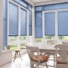 Blue horizontal Venetian blinds covering tall white-framed bay windows and door, lowered and filtering daylight; dining table and chairs in a sunlit kitchen/dining nook with cushions and garden view.