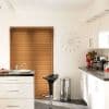 Brown wooden horizontal blinds lowered over the window, closed and filtering daylight into a modern white kitchen with black counters, sink, bar stool, stainless bin, wall clock and red tulips.