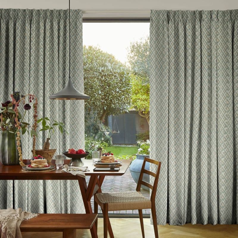 Patterned pleated floor-to-ceiling curtains, drawn apart at the center to frame the opening, revealing sunlight and a leafy backyard; wooden dining table set with breakfast, pendant light overhead.