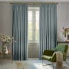 Pleated blue-gray curtains on a brass rod, hung full-length and parted at center, filtering daylight and framing a paneled French window in a softly lit living room with green armchair.