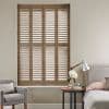 Full-height wooden plantation shutters with four panels and louvers, closed with slats tilted to filter daylight; set in a neutral-toned bedroom beside a tufted armchair, bed, and bedside table lamp.