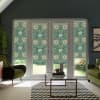 Patterned floral roller blinds hang lowered over glass patio doors, filtering sunlight into a contemporary living room with olive-green armchairs, glass coffee table, zigzag rug, and houseplants.