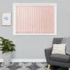 Vertical pink fabric blinds cover a rectangular window, closed and lightly filtering daylight; set on a gray living-room wall with a gray armchair, patterned rug, potted plant, and framed art.