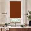 Brown roller blind covers most of a kitchen window above a stainless sink, complementing a countertop with wooden cabinets, a bowl of greens, potted plants, and a framed sketch on the wall.