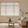 White horizontal blinds covering a large rectangular window, fully lowered with slats closed, softly filtering daylight; in a neutral living room with beige walls, sofa, wooden coffee table and lamp.
