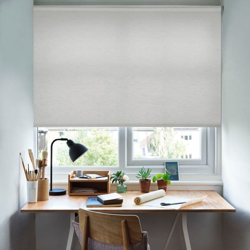 A white fabric roller blind pulled down over the window, filtering daylight; a wooden desk beneath with lamp, art brushes, notebooks and potted succulents, bright suburban view outside.