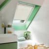 Cream pleated blind on a skylight, partially lowered and filtering daylight; green-painted sloped window frame and white herringbone-tiled bathroom with sink, bathtub, plant, and bath tray.