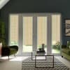 French doors with multicolored vertical-striped blinds cover patio windows, letting soft light into a living room furnished with green armchairs, a glass coffee table, patterned rug, and potted plants.