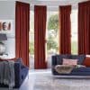 Rust-red pleated floor-length curtains across a bay window, partially closed and filtering sunlight; modern living room with navy sofas, patterned rug, lamp, and a leafy garden visible outside.