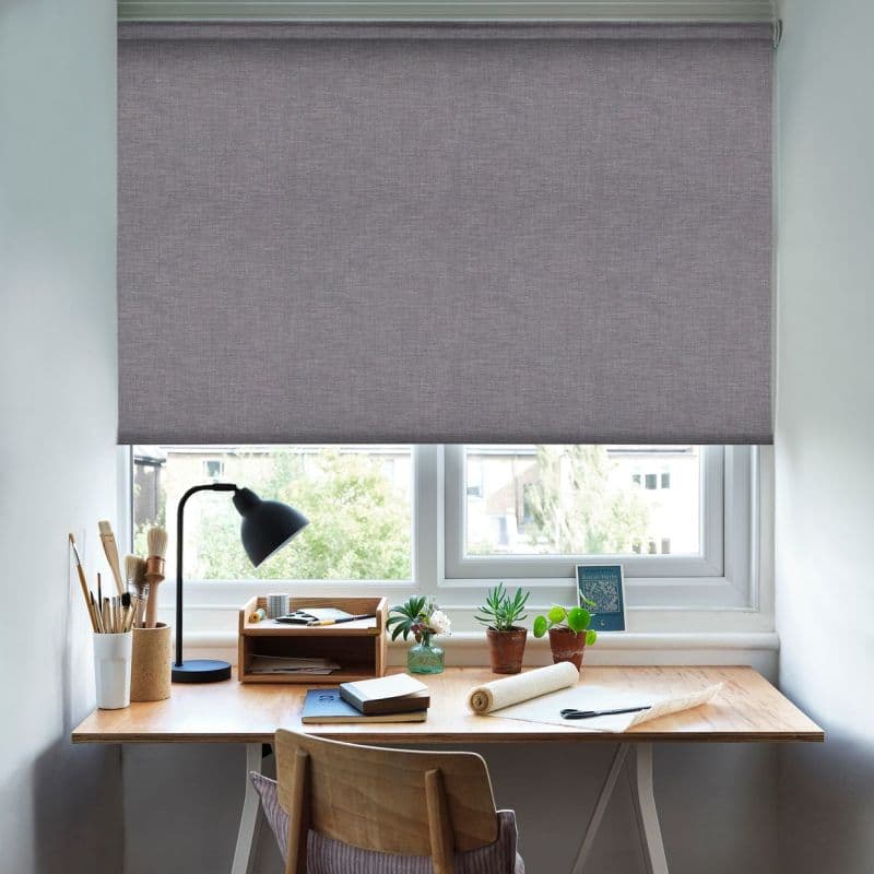 A grey textured roller blind lowered over a double window, blocking light; beneath it a wooden desk workspace with a lamp, books, potted plants, brushes and a chair in daylight.