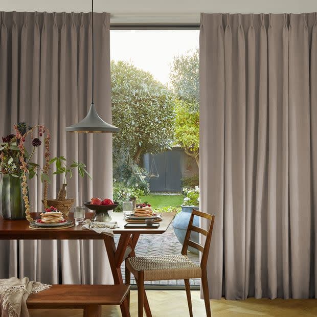 Floor‑length pleated beige curtains, drawn to the sides and framing a glass door, letting daylight in; wooden dining table set for breakfast with pendant lamp, chairs, and backyard garden beyond.