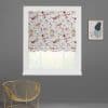 A white Roman blind with pink-red floral and green leaves, partially lowered and softly folded, filtering daylight through a white-framed window; grey wall, gold chair, abstract print.