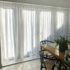 Sheer white, floor-length pleated curtains drawn closed on a wide patio door, filtering bright sunlight into a tiled dining area with a wooden table, black chairs, and a potted plant.