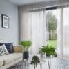 Sheer beige, ceiling-mounted curtains—full-height with soft vertical folds—slightly parted, filtering daylight; in a bright living room beside a pale sofa and patterned tile floor, potted plants foreground, garden visible.