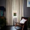 Cream full-length curtains with sheen flank a small window; a Roman blind is partially lowered, filtering daylight into a dim modern living room with dark floor and a woven lounge chair.