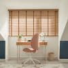 Wooden horizontal blinds covering a wide dormer window, slats tilted to filter sunlight; a compact desk with lamp, plant and pink office chair in a bright attic study.