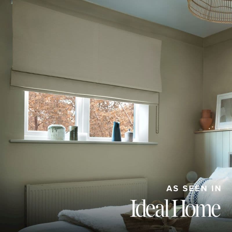 Beige Roman fabric blind, wall‑mounted and partially lowered with a beaded pull, filtering soft daylight over a small bedroom window; sill holds vases, view of brown autumn trees.
AS SEEN IN IdealHome