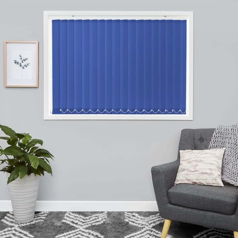 Blue vertical blinds, fully closed with bead chains at bottom, covering a white-framed window; filtering light into a gray living room with a gray armchair, cushion, knit throw, and potted plant.