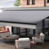 Large gray retractable awning with straight valance, fully extended over sliding glass doors, shading a modern outdoor patio with wooden deck, table, cushioned seating and brick facade in daylight.