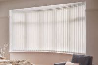 Vertical fabric blinds across a curved bay window, fully closed and diffusing soft daylight; set in a softly lit bedroom corner with a bed, bedside lamp and upholstered chair.