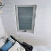 Pleated skylight blind, fully lowered in a centered white-framed roof window with blue-grey horizontal stripes; filtering soft daylight in a white-paneled attic bedroom with bed, pillows and guitar-shaped cushion.