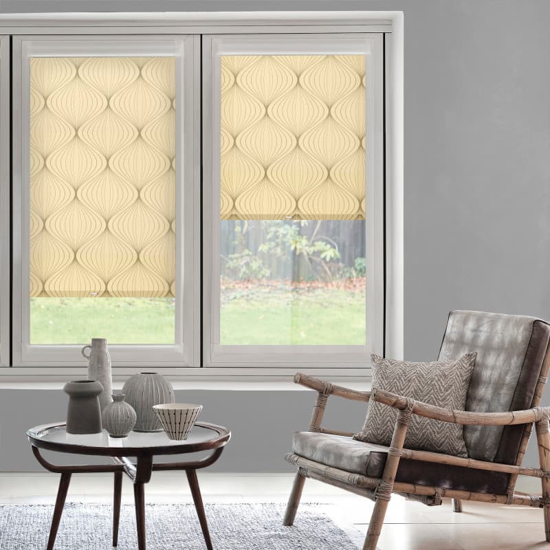 Beige roller blinds with curved pattern — left lowered, right partially lowered, filtering daylight into a gray living room with rattan armchair, cushion and round table with vases, grassy backyard view.
