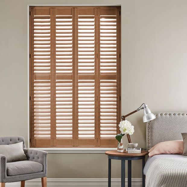 Wooden four-panel plantation shutters with horizontal louvers, closed and slightly angled to filter daylight, covering a tall window in a neutral bedroom corner—bed, bedside table, lamp and armchair.