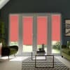 Glass patio doors with coral-red roller blinds, partially lowered to filter sunlight into a modern living room with green armchairs, a glass coffee table on a chevron rug, and a garden beyond.