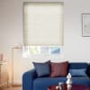 Cream cellular (honeycomb) blind lowered over window, fine horizontal pleats filtering soft daylight; above a window showing roof tiles and trees, in a modern living room with a blue sofa.