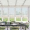 Dining table set with four stacked plates, small candles and green glassware, surrounded by green cushioned chairs in a sunlit conservatory with large windows overlooking a grassy backyard and fence.