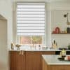 A white zebra roller blind, lowered with a small bottom gap, alternating sheer and opaque bands filtering daylight; kitchen with marble counters, oak cabinets and brass faucet, brick courtyard outside.