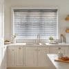 White horizontal Venetian blinds fully covering the window, slats slightly tilted, filtering soft daylight; above a stainless sink on a white marble countertop with cream cabinets, potted herb and utensils.