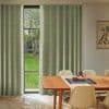 Green patterned curtains, floor-length on each side of a tall three-pane window, hanging in soft folds, partially open and filtering daylight; dining room with wooden table, chairs, garden view.