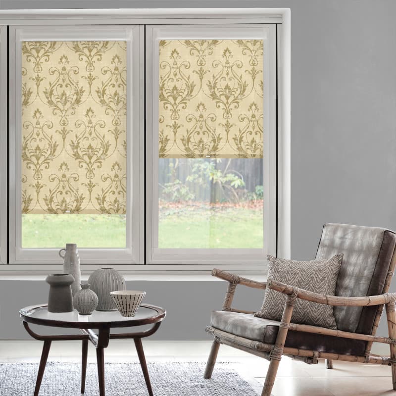 Patterned beige roller blinds with damask motif lowered over a double window; partially closed, filtering daylight; cozy living room with rattan armchair, coffee table with ceramics, grassy backyard visible.