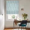 A pale teal Roman blind with white feather motif, lowered and softly folded, filters daylight over a small desk and teal chair in a bright, neutral home study with a vase of flowers.