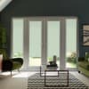 Glass double doors with lowered pale roller blinds filter daylight into a cozy living room; a glass coffee table sits on a chevron rug between green upholstered chairs and plants.