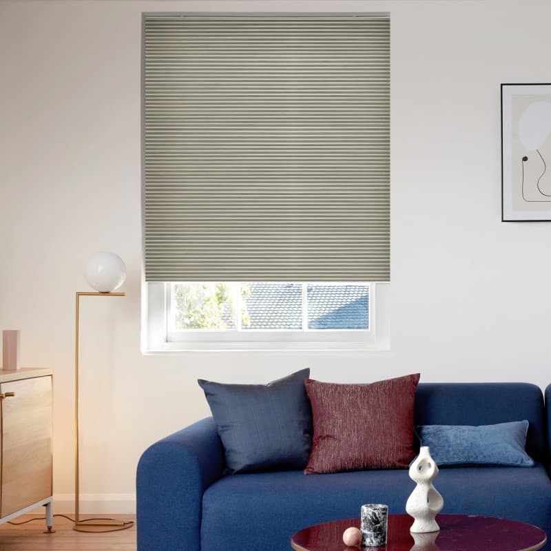 Beige narrow‑pleated blind, lowered to cover most of the window with a bottom gap, filtering daylight. Bright living room with blue sofa, cushions, floor lamp and framed art; rooftop beyond.