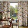 Pleated full-length floral curtains (white with yellow‑orange flowers) are parted to reveal a sliding glass door, filtering soft daylight over a breakfast table and wooden chairs overlooking a sunlit garden.