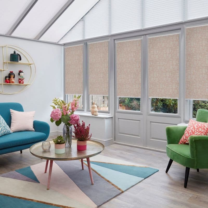 Beige patterned roller blinds cover tall conservatory doors and lower windows, partially closed to filter daylight, in a bright, pastel-furnished sunroom with colorful sofa, rug, and plants.