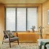 Horizontal layered roller blinds over three tall windows, alternating sheer and opaque stripes, partially closed filtering bright daylight; warm wood-paneled sitting nook with armchair, side table, plant, rug.